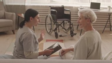 Elinde dijital tabletle, arka planda tekerlekli sandalyeyle evdeki yumuşak koltukta oturan mutlu yaşlı kadınla konuşan genç bayan sosyal hizmet görevlisinin orta ölçekli bir fotoğrafı.