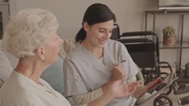 Gülümseyen genç bayan sosyal hizmetler görevlisinin, dijital tableti nasıl kullanacağını söylerken, rahat bir dairede, kanepede yan yana otururken. Arka planda duran tekerlekli sandalye