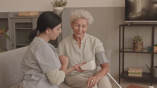 坐在舒适公寓沙发上的年轻女性社会工作者测量老年妇女血压的中刺镜头 — 图库视频影像