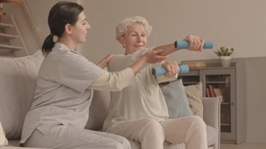 Genç bayan sosyal görevlinin, gülümseyen yaşlı kadının fiziksel egzersiz yapmasına yardım ederken, rahat bir dairede kanepede yan yana otururken orta boy fotoğrafı.