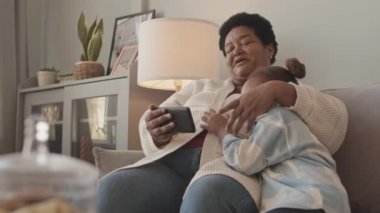 Orta uzunluktaki Afro-Amerikan kadın ve sevimli küçük torunun gündüz vakti evdeki koltukta oturup gülümsemesi, konuşması ve akıllı telefondan video izlemesi.