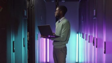 Neon ışıkla aydınlatılmış raflarda duran ve bilgisayarla çalışmalarını analiz eden Afrikalı genç bir bilişim yöneticisinin orta boy görüntüsü.