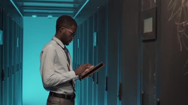 若いアフリカ系アメリカ人の男性の中程度のショットは ラックサーバーキャビネットに立って デジタルタブレット上で作業を着て — ストック動画