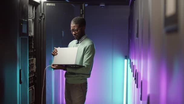 若いアフリカ系アメリカ人男性のIt技術者のロックダウンノートパソコンを使用してネオンライトで照らされたデータセンターでのハードウェアの動作をチェック — ストック動画
