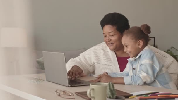 一位快乐的五岁小女孩 白天坐在客厅的办公桌前 微笑着 使用手提电脑 坐在非洲裔美国人祖母的膝上 进行中度特写 — 图库视频影像