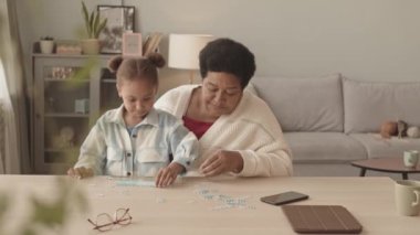 Sevimli beş yaşında bir kızın belinde neşeli Afrika kökenli Amerikalı bir büyükannenin dizlerinin üzerinde oturmuş gündüz vakti masasında konuşuyor, gülüyor ve bulmaca topluyor.