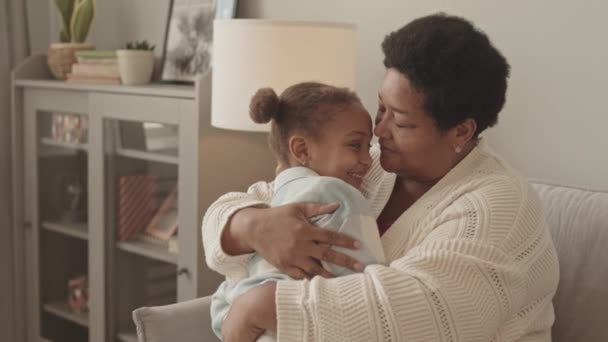 中等程度的非洲裔美国成年女性坐在沙发上 白天在客厅里笑着拥抱可爱的5岁小女孩 — 图库视频影像