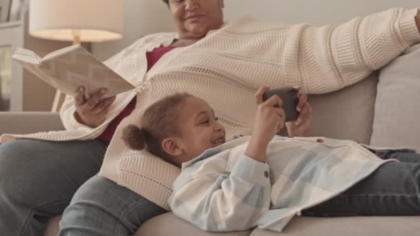 中等长度的非洲裔美国成年女性和可爱的5岁女孩白天在客厅的沙发上休息 孩子们用智能手机 奶奶看书 — 图库视频影像