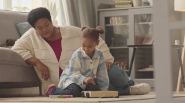 Düşük açılı, mutlu beş yaşında bir kız, evinde oturma odasında oturmuş renkli ahşap oyuncaklarla oynuyor, Afrikalı Amerikalı büyükanneyi seviyor, onu denetliyor, konuşuyor ve gülümsüyor.