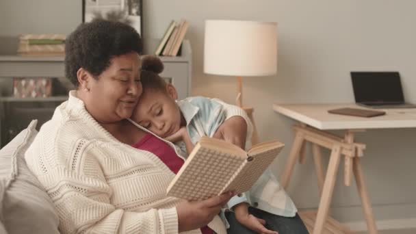 楽しいです5歳の女の子の膝の上に座っています彼女のお気に入りのアフリカ系アメリカ人の祖母誰が昼間のリビングルームで声を出して本を読んで — ストック動画
