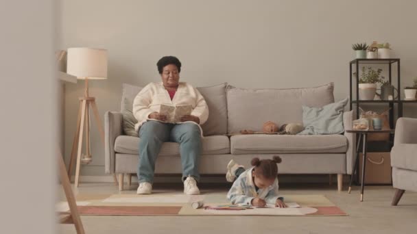 一大片可爱的小女孩躺在地板上 用铅笔在纸上画画 然后站起来和非洲裔美国奶奶坐在沙发上 白天在客厅看书 — 图库视频影像