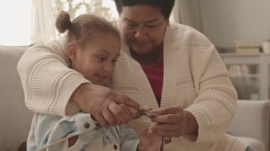 Orta boy Afrika kökenli yetişkin bir Amerikalı kadının gündüz vakti oturma odasında kanepede oturup beş yaşındaki torununa örgü örmesinde yardım etmesi.