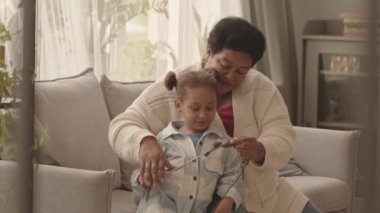 Orta uzunluktaki Afro-Amerikan kadın gündüz vakti kanepede oturuyor ve beş yaşındaki torununa örgü örmeyi öğretiyor.