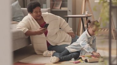 Beş yaşında mutlu bir kız çocuğu, evinde oturma odasında oturmuş renkli tahta oyuncaklarla oynuyor, Afro-Amerikan büyükannesini seviyor, onu akıllı telefonuyla çekiyor, konuşuyor ve gülümsüyor.