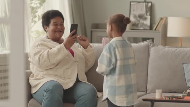 中年快乐的非洲裔美国老年妇女在白天坐在客厅的沙发上 手持智能手机 拍摄着她的小孙子 她的小孙子正在吹毛求疵 — 图库视频影像