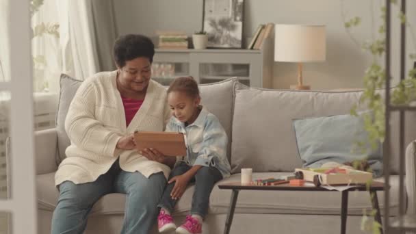 白天黑夜 快乐的非洲裔美国老年妇女坐在客厅的沙发上 与可爱的五岁小女孩一起使用平板电脑 她们谈笑风生 — 图库视频影像