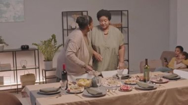 Orta uzunlukta iki genç ve yaşlı Afro-Amerikan kadının bayram kutlaması için sofrayı kurması, çocukların arka planda koltukta oturması.