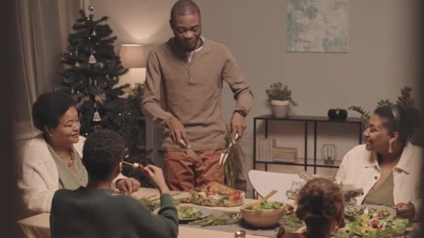 中等程度快乐的黑人年轻人在家人坐着庆祝圣诞节的桌上切烤鸡 — 图库视频影像