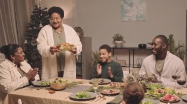 Orta uzunluktaki kıdemli Afrikalı Amerikalı kadın aile üyelerine kızarmış tavuk ikram ederek evde yemek masasında Noel 'i kutluyor.