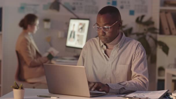 彼の女性の同僚のビデオ会議を背景にしながら 夜にオフィスの机の上に座ってノートパソコンを入力眼鏡で若いアフリカ系アメリカ人男性のオフィスワーカーのウエストアップショット — ストック動画