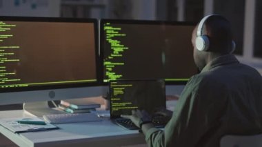 İki monitör ve dizüstü bilgisayar program kodları önünde oturan, boş bir ofiste geç saatlere kadar çalışan, kulaklıklı Afro-Amerikalı programcının dikiz görüntüsü.