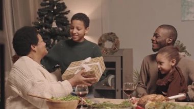 Orta boylu, kısa saçlı siyahi kız Noel hediyesini aile arasında şenlik masasında oturan, sarılan ve torununu öpen favori büyükannesine veriyor.