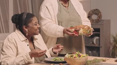 Orta uzunlukta kesilmiş bir kadın, evde yemek masasında oturan, Noel 'i kutlayan, el çırpan siyahi aile üyelerine kızarmış tavuk servis ediyor.