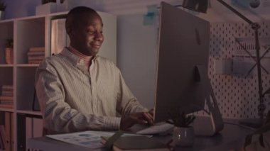 Orta yaşlı Afro-Amerikan bir adamın yavaş çekimleri klavyede yazarken gülümsemesi, ofiste bilgisayar monitörünün önünde oturması, gece geç saatlere kadar tek başına çalışması.