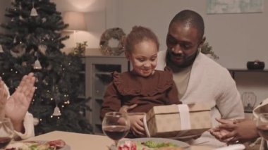 Mutlu siyah kadın şenlik masasında oturuyor, küçük tatlı kızını şaşırtıyor, genç babasının kucağında Noel hediyesiyle oturuyor, kocası karısına hediye veriyor.