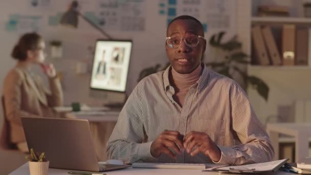 中Povショットの若いアフリカ系アメリカ人男性の眼鏡で暗いオフィスの机に座ってビジネス文書を議論するカメラを見て 背景に彼の秘書ビデオ会議ながら — ストック動画