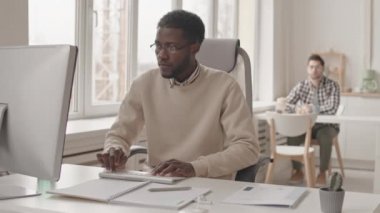 İş arkadaşı arka planda öğle yemeği molası verirken, modern ofiste bilgisayar üzerinde çalışan Afro-Amerikan bir adamın yavaş çekimleri.