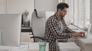 Sakallı, gözlüklü, dizüstü bilgisayarda çalışan ve ofisteki iş yerinde oturan melez bir programcının fotoğrafı.