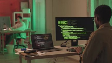 İş arkadaşıyla birlikte büroda kırmızı ve yeşil ışıklandırmayla birlikte bilgisayar başında oturan Afrikalı-Amerikalı bilişim uzmanı hackleme yazılımının dikiz görüntüsü. Siyah ekranda çalışan program kodu