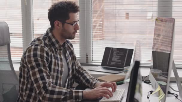 髭を生やした混合レースプログラマーのサイドビュー メディア ショット現代のオフィスの窓のそばに座ってコンピュータ上でカジュアルウェアや眼鏡書き込みプログラムコードで — ストック動画