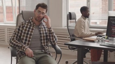Bilgisayar üzerinde çalışan Afrikalı-Amerikalı meslektaşı modern ofiste kalırken yazılım geliştiricisinin akıllı telefondan konuşmasının yavaş çekimleri.