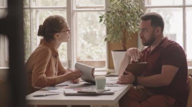 Modern tasarım ajansında masa başında oturan müşteriye planlar gösteren genç bayan tasarımcının orta yavaş çekimi.