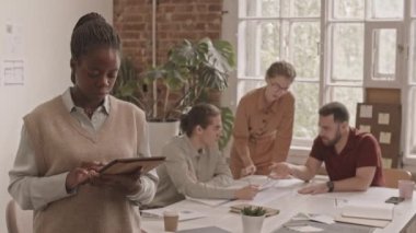Modern tasarım ajansında dijital tabletle kameraya poz veren genç Afro-Amerikalı kadın tasarımcının orta yavaş portresi. Meslektaşları arka planda proje üzerinde çalışıyor.