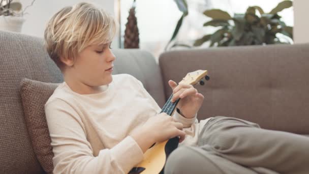 中等长度的短发白人少女坐在沙发上 慢慢地在家里弹奏四弦琴 — 图库视频影像