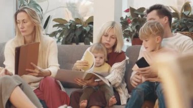 Orta uzunlukta beyaz bir aile gündüz vakti oturma odasında oturuyor, konuşuyor, tablet bilgisayar kullanıyor, defterlere bakıyor, gülümsüyor.