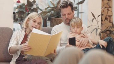 Orta uzunlukta beyaz bir adam çocuklarla birlikte oturma odasında oturuyor, akıllı telefon kullanıyor ve çocuklara gösteriyor.