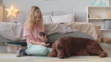 8 yaşındaki güzel bir kızın akıllı telefonuyla şirin İrlandalı köpeğini çekerken ve yerde, yatak odasının yanında otururken yavaş çekimde çekilmiş.