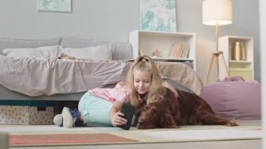 Küçük bir kızın, şirin İrlanda sosisli köpeğinin kucak kucağa selfie çekerken ve akıllı telefonunda rahat yatak odasında yerde yan yana yatarken çekilen yavaş bir fotoğraf.