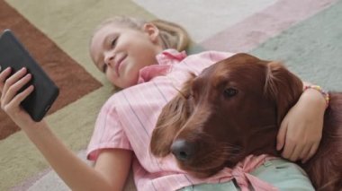 Yukarıdan, 8 yaşındaki gülümseyen kızın yatak odasında tatlı İrlandalı köpeğiyle birlikte yatarken akıllı telefondan kayışının orta ölçekli görüntüsü.