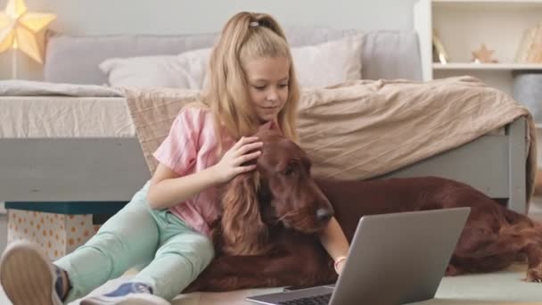 Slowmoショットの幸せな8歳の女の子座っています床に居心地の良いベッドルームで一緒に彼女のかわいいですアイルランドのセッター犬ながらビデオチャットの友人とラップトップ — ストック動画