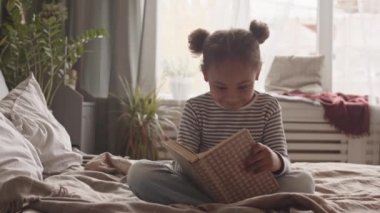 5 yaşında Afro-Amerikan bir kızın, rahat bir yatak odasında, yatakta oturup komik kitaplar okumasının yavaş çekimleri.