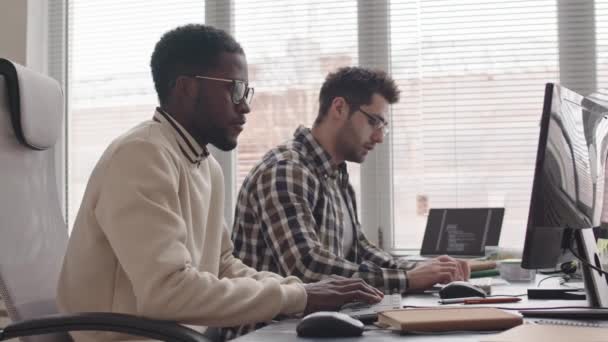 サイドビューのメディアは 現代のオフィスで窓のそばに座っている新しいソフトウェアを開発する2人の若い多民族男性プログラマーの遅いショット — ストック動画