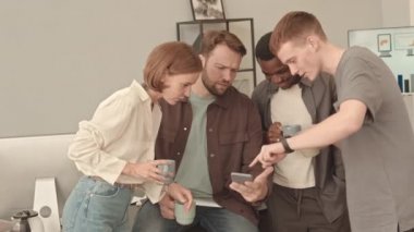 Orta uzunlukta, dört mutlu genç meslektaşın gündüz vakti ofise girip kahve içmesi, akıllı telefon kullanması ve gülümsemesi.