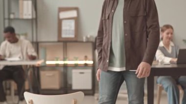 Kafkasyalı sakallı genç bir adamın kareye girmesi, normal bir iş gününde koltukta sırtüstü oturması, kameraya bakıp gülümsemesi.