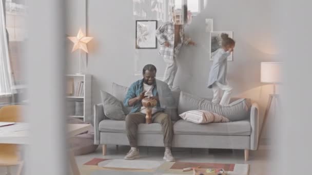 两个有趣的小女孩在沙发上跳着 而她们的父亲在旁边弹奏着非洲鼓声 坐在舒适的家里玩乐 — 图库视频影像