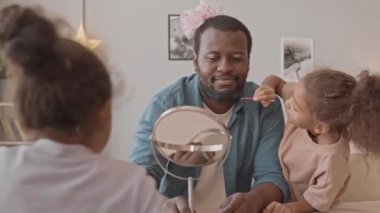 Kafasında pembe kurdele olan Afro-Amerikan bir adamın, iki güzel kızı eğlenmek için yüzüne makyaj yaparken, aynaya baktığı orta yavaş bir fotoğraf.